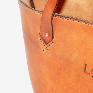 cropped image of a caramel brown leather tote bag with hand stitched detailing and metal hardware.
