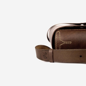 side view of a a rich brown leather satchel with shoulder strap, etched logo and hand stitched detailing.