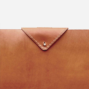 cropped image of a camel brown leather laptop sleeve featuring a triangle shaped leather closure with white stitching and brass hardware detailing.