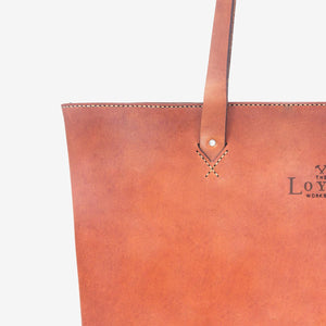 Cropped image of a caramel brown leather tote bag featuring hand stitched detailing and an etched logo imprinted in the leather.