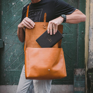 man in a street taking a black notebook out of a brown leather messenger bag he is wearing crossbody