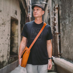image of a man standing in a concrete alleyway wearing a black tshirt and cap and a caramel brown leather messenger style bag worn cross body.